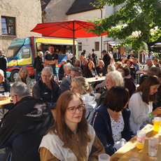 Draußen sitzen viele Menschen an Holzbänken und Tischen, genießen Getränke in Plastikbechern und essen. Im Hintergrund ist ein Food-Truck sowie ein gemütliches Gebäude mit Schildern zu sehen. Die Atmosphäre wirkt gesellig und lebhaft.