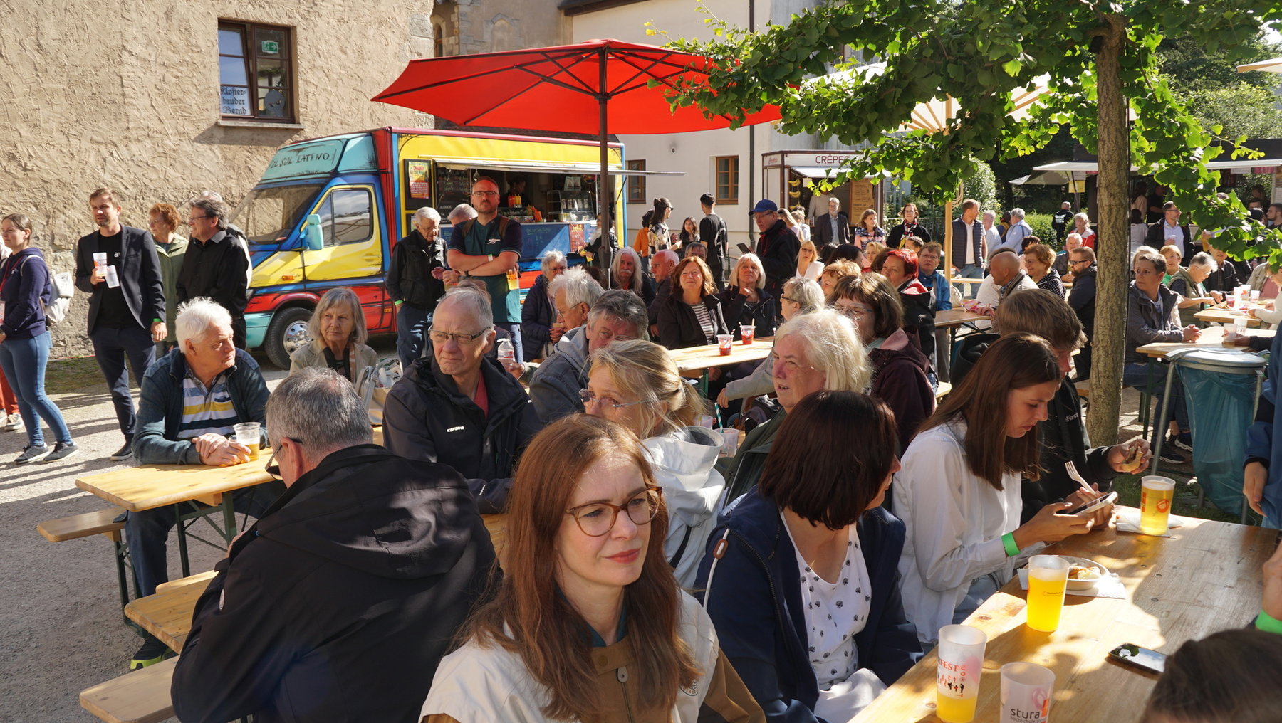 Draußen sitzen viele Menschen an Holzbänken und Tischen, genießen Getränke in Plastikbechern und essen. Im Hintergrund ist ein Food-Truck sowie ein gemütliches Gebäude mit Schildern zu sehen. Die Atmosphäre wirkt gesellig und lebhaft.