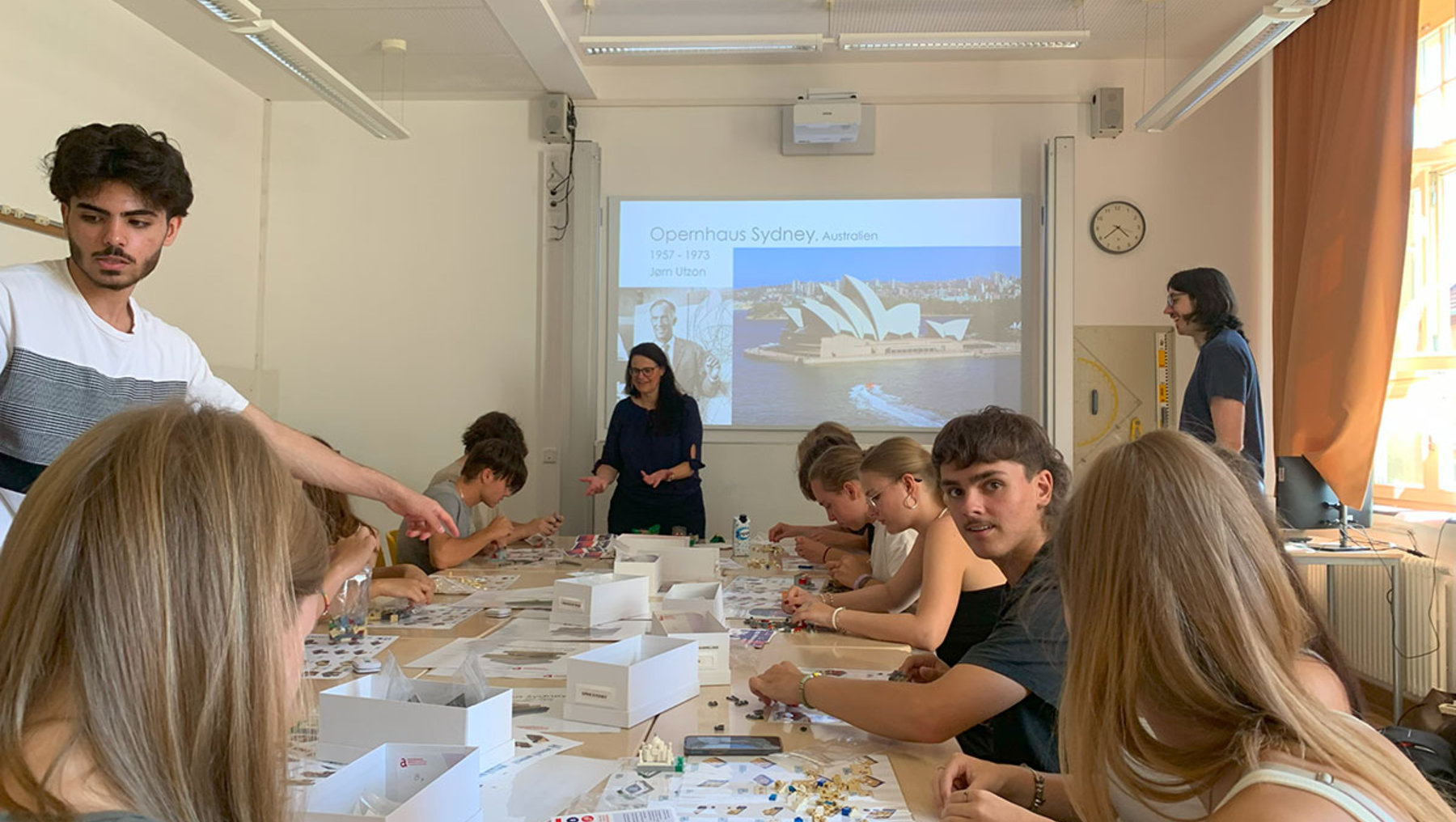 Ein Workshop-Raum mit mehreren Teilnehmenden, die an langen Tischen sitzen und kreativ mit Materialien wie Papier, Kleber und Bildern arbeiten. Eine Person steht vorne und erklärt etwas, während ein Bildschirm im Hintergrund eine Präsentation mit dem Titel „Greenhead-Sydney-Australia“ zeigt.