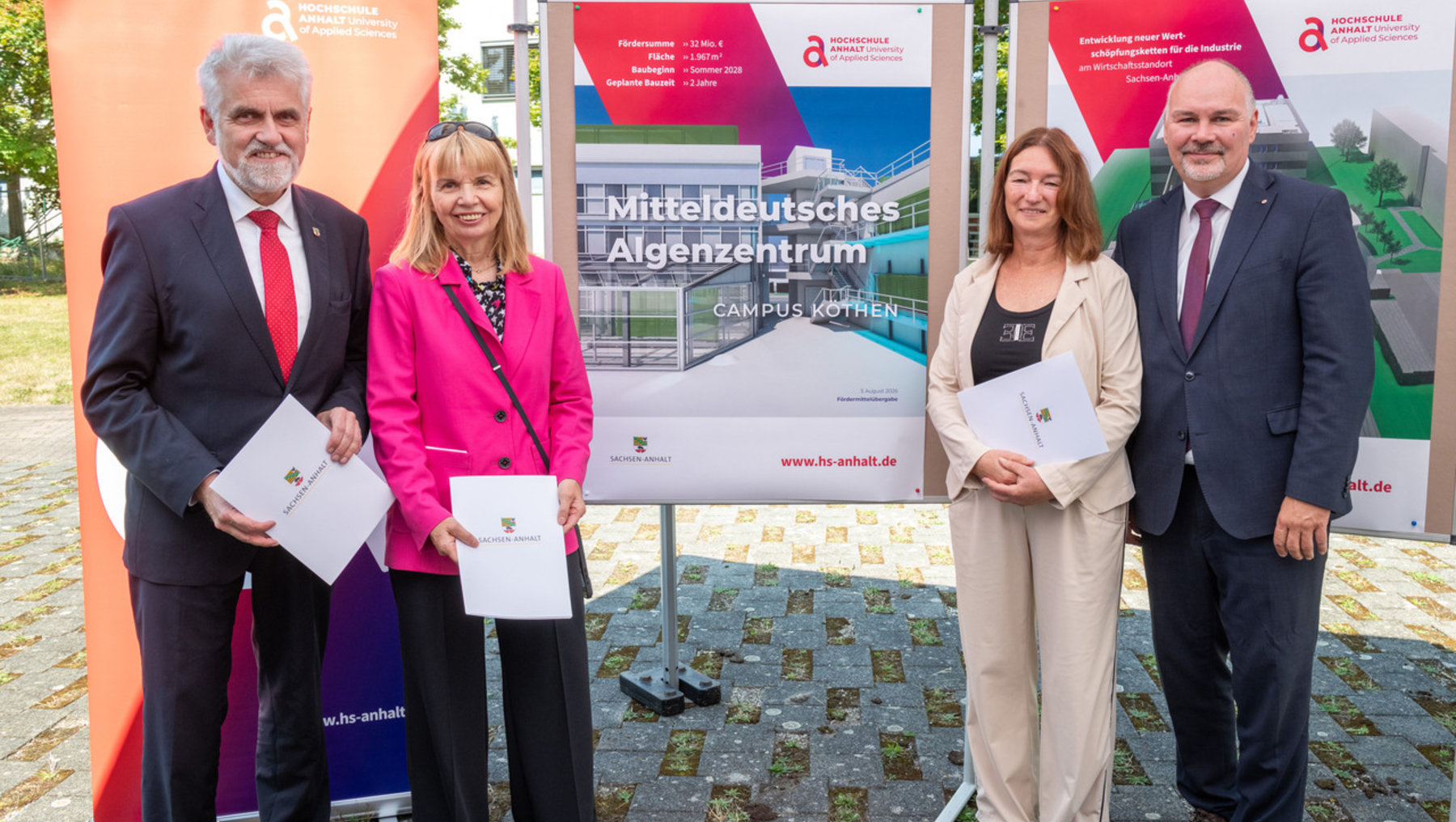 Vier Personen stehen vor einem Plakat zur Eröffnung des Mitteldeutschen Algenzentrums am Campus Kothen der Hochschule Anhalt. Sie halten Urkunden und lächeln in die Kamera.
