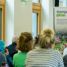 Gruppe von Menschen in einem Raum, die sich einem Plakat mit dem Titel Kompetenz Grün zuwendet. Das Plakat zeigt eine grüne Landschaft mit Pflanzen und bezieht sich auf Kompetenzzentren und Transformation im Bereich Grünflächen und Nachhaltigkeit.