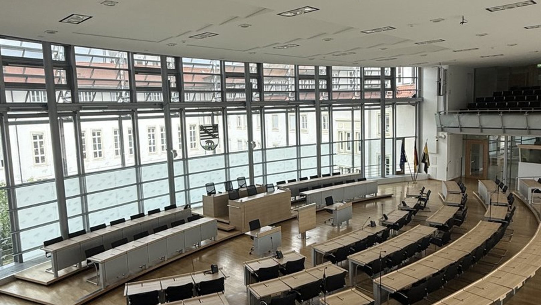 Großer, leerer Hörsaal mit halbkreisförmig angeordneten Sitzreihen aus Holz und Metall, zentralem Podium mit Laptop-Arbeitsplatz, großen Fenstern an der Stirnseite und indirekter Deckenbeleuchtung.
