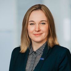 Eine lächelnde Person mit mittellangem, hellbraunem Haar, die ein dunkles Sakko über einem karierten Rollkragenpullover trägt und professionell vor neutralem Hintergrund posiert.