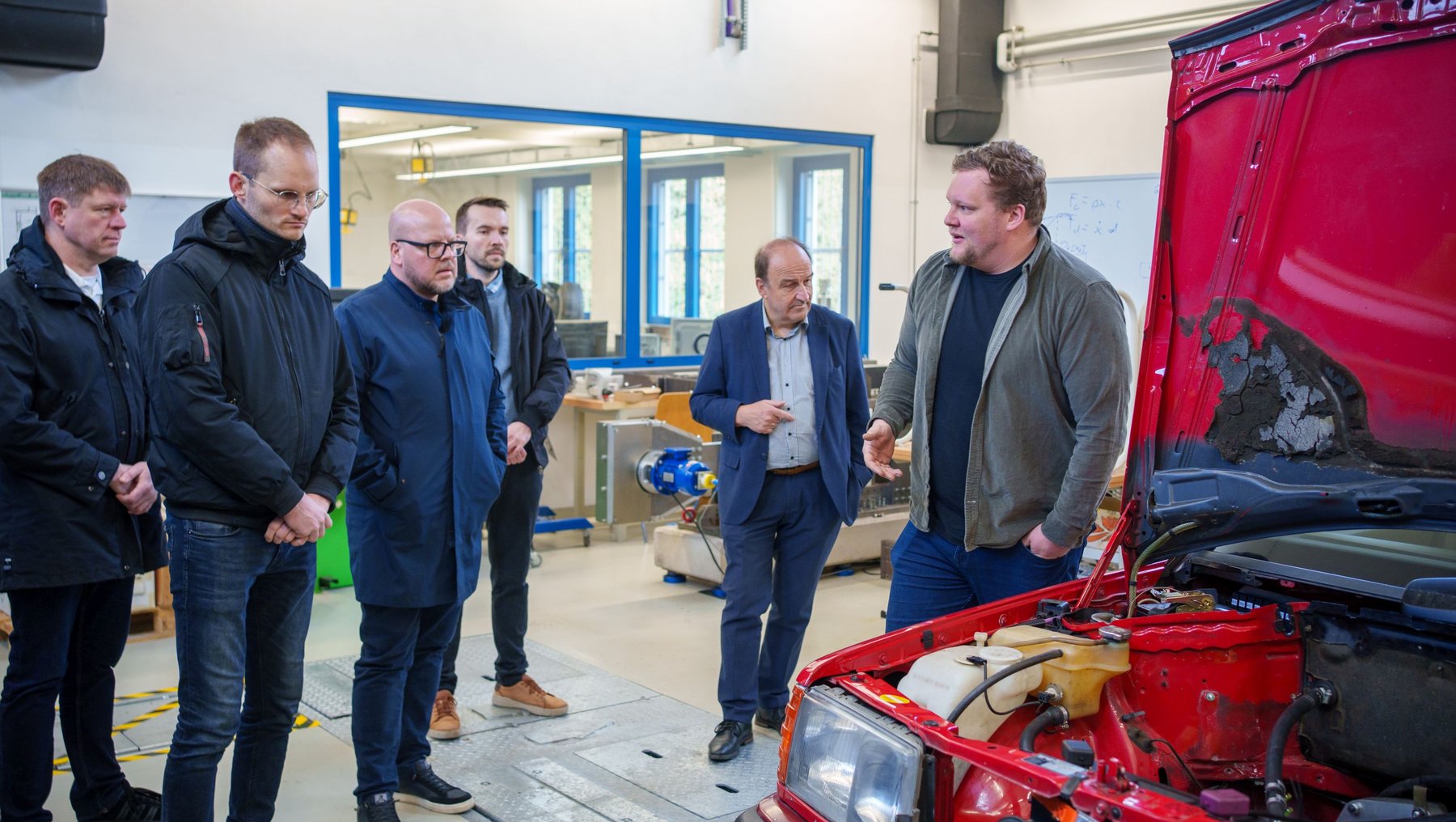 Fünf Teilnehmer in der Fahrzeugtechnik die sich um ein offenes rotes Fahrzeug mit geöffneter Motorhaube versammelt haben. Einer erklärt mit Gesten etwas über den Motor, während die anderen aufmerksam zuhören.