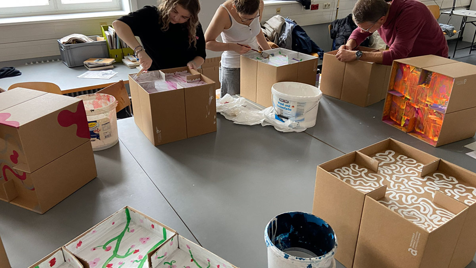 Drei Personen bemalen gemeinsam mehrere Kartons mit bunten Mustern und Motiven wie Herzen und Wirbeln. Im Raum liegen weitere bemalte Kartons, Eimer mit Farbe und Pinsel.
