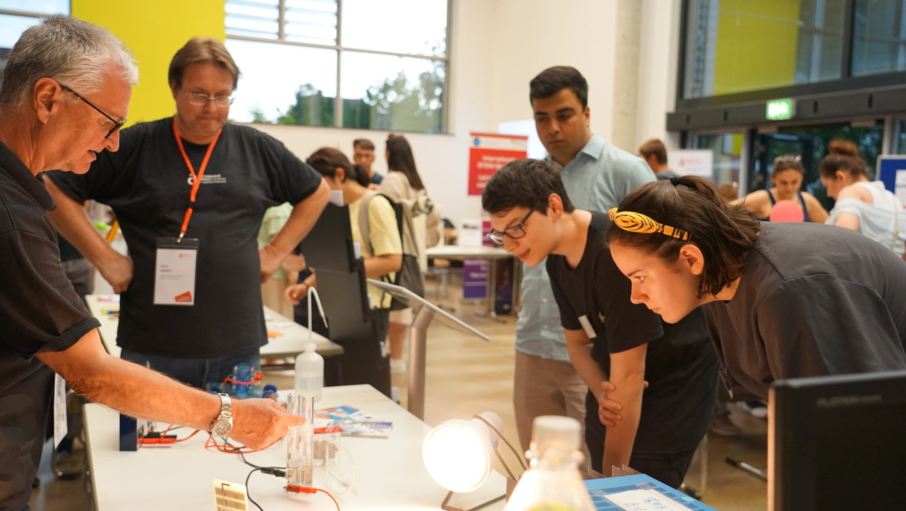 Eine Gruppe von Menschen, darunter Erwachsene und Jugendliche, beobachtet und experimentiert an einem Tisch mit elektronischen Bauteilen und einer Schaltung, die eine kleine Lampe mit Strom versorgt.