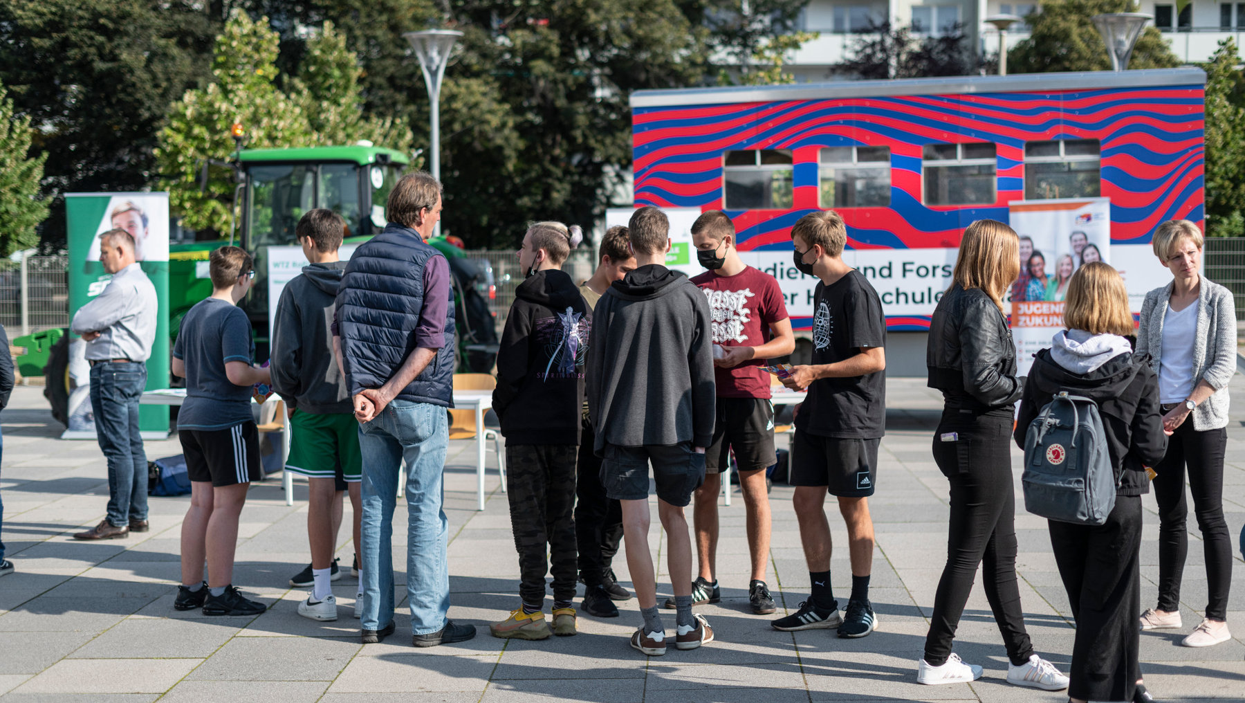 Gruppe junger Menschen steht im Freien und wartet vor einem roten Bus mit Aufschrift „Bildung und Forschung“.