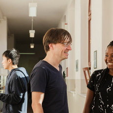 A group of six people, including men and women, are walking and talking in a hallway. They appear to be in a good mood, smiling and engaging with each other. The hallway has large windows and a few plants. The setting seems to be indoors, possibly in an office or educational building.