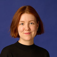 Porträt einer Person mit roten, mittellangen Haaren, die freundlich lächelt. Sie trägt ein schwarzes Oberteil und hat kleine Ohrringe.