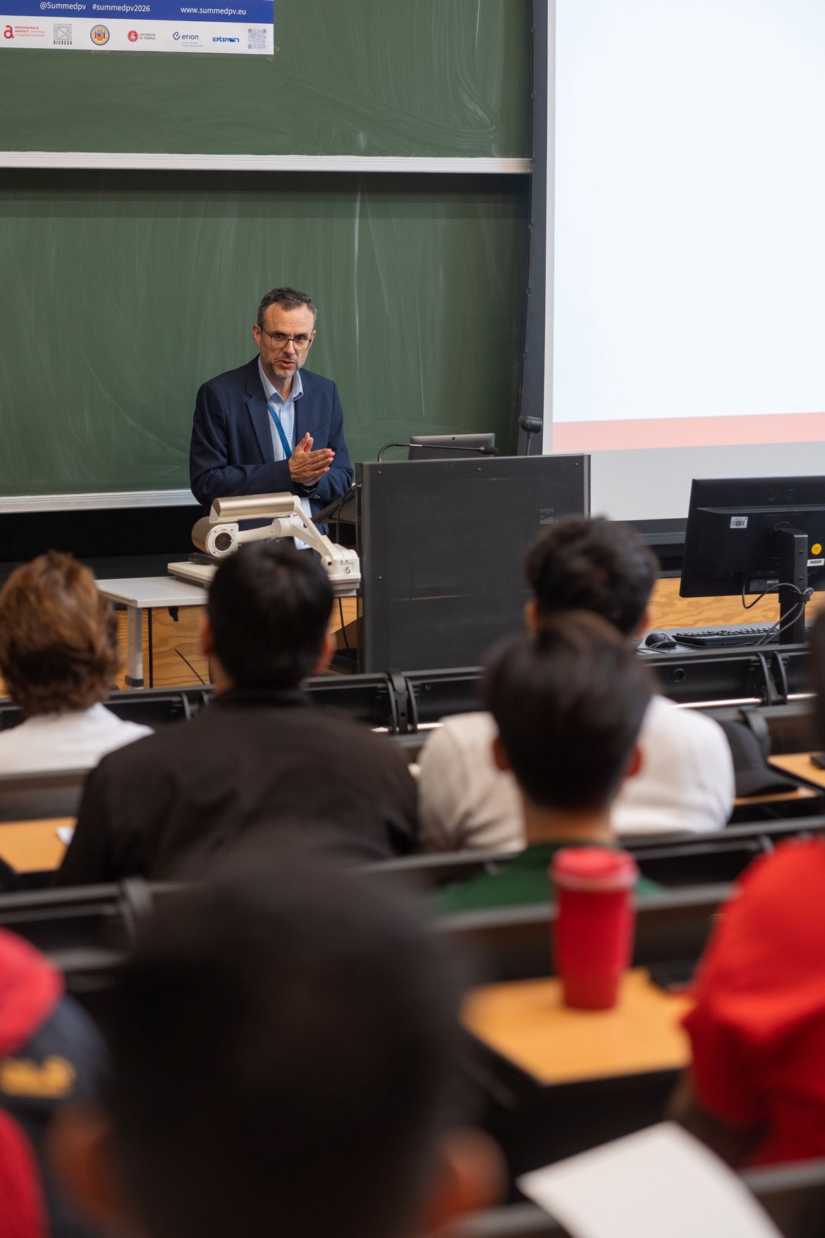 Ein Vortragender steht vor einem Publikum in einem Hörsaal. Er hält eine Präsentation und steht hinter einem Pult mit Technik. Die Zuhörer sitzen auf Stühlen, einige mit Getränken, und blicken aufmerksam in seine Richtung.