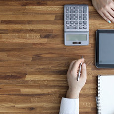Two hands working at a wooden desk with a tablet displaying a document, a calculator, a smartphone, a blue notebook, a white notebook with a pen, a clipboard with charts, and a pair of glasses.