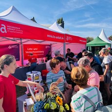 Ein Stand der Hochschule Anhalt mit dem Schwerpunkt Lehr- und Versuchsweinberg. Eine Mitarbeiterin in rotem Shirt reicht einer Besuchergruppe Proben von Trauben und Weinblättern. Im Hintergrund weitere Interessierte und ein roter Zeltaufbau mit Logo und Webadresse.
