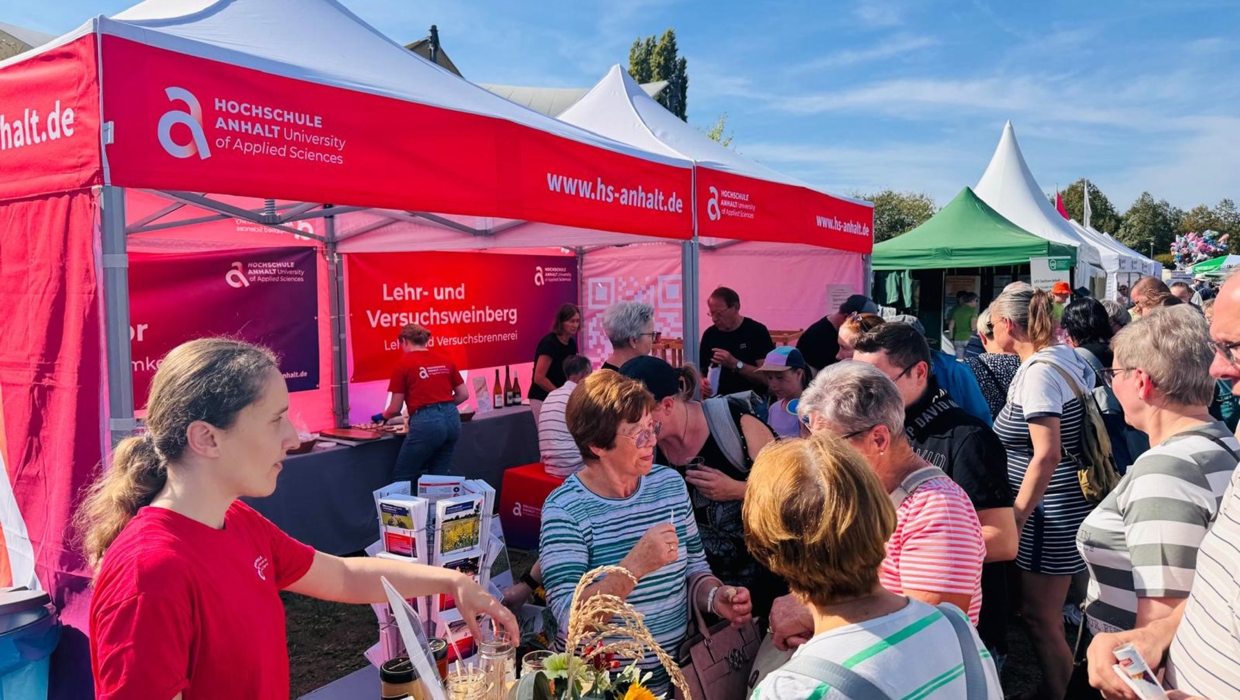 Ein Stand der Hochschule Anhalt mit dem Schwerpunkt Lehr- und Versuchsweinberg. Eine Mitarbeiterin in rotem Shirt reicht einer Besuchergruppe Proben von Trauben und Weinblättern. Im Hintergrund weitere Interessierte und ein roter Zeltaufbau mit Logo und Webadresse.