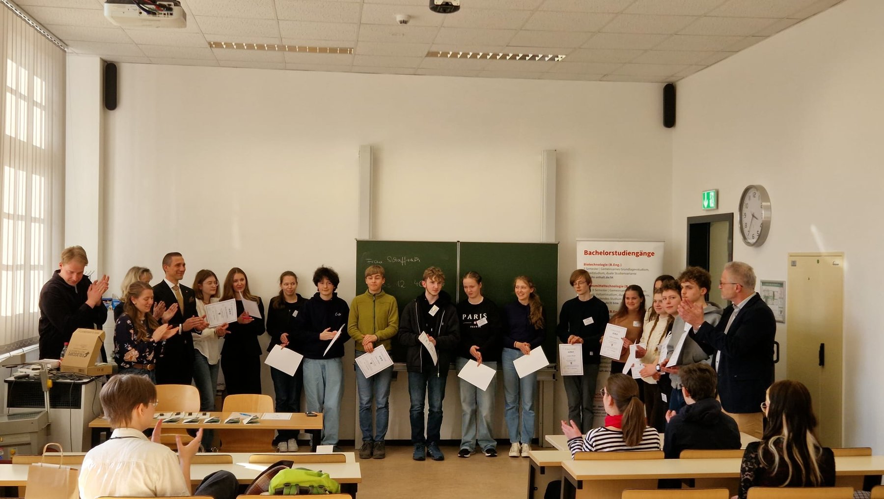 Gruppe von Schülern und Erwachsenen steht im Klassenzimmer, hält Urkunden oder Zertifikate und wird von zwei Personen mit Applaus gefeiert.
