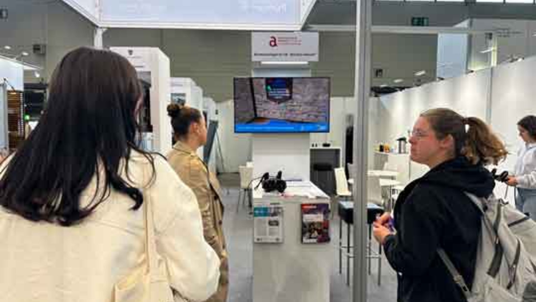 Drei Personen auf einer Messe: Eine Frau mit langem Haar von hinten, eine Person in Uniform und eine Frau mit Rucksack, die sich vor einem Messestand mit Bildschirm und Informationsmaterial unterhalten.