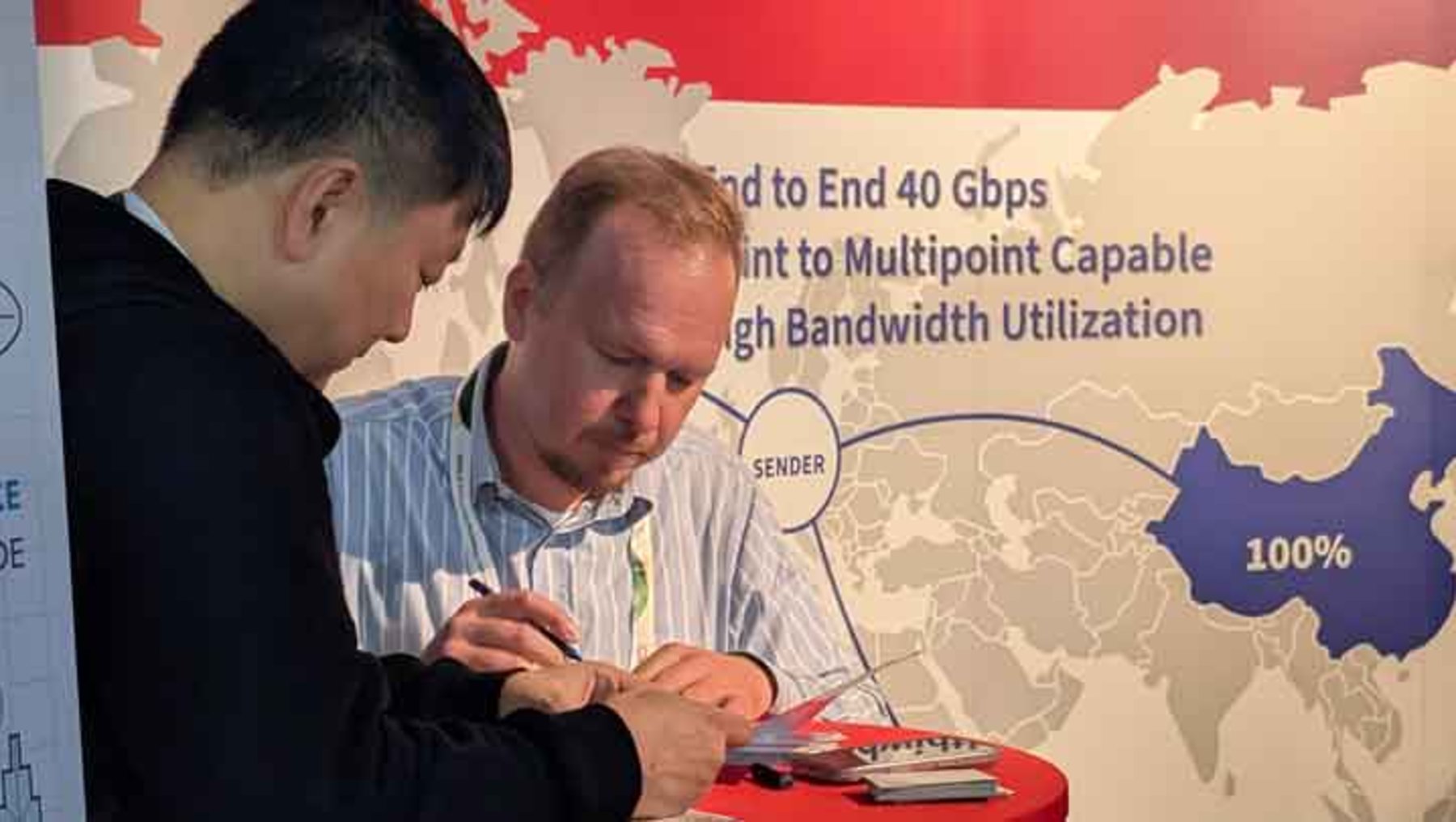 Zwei Männer unterschreiben gemeinsam ein Dokument vor einem Infoposter mit einer Weltkarte, die China und globale Datenübertragung mit bis zu 40 Gbps und 100-prozentiger Bandbreitennutzung zeigt.