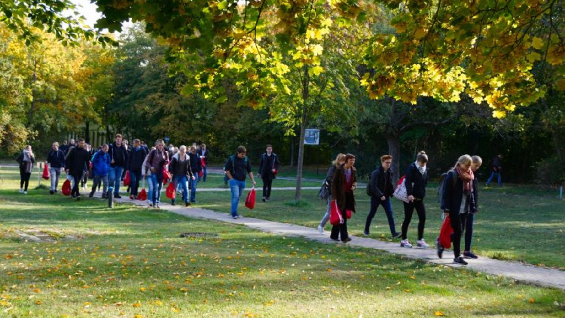 Eine Gruppe von Menschen geht auf einem Weg durch einen Park. Die Bäume haben bunte Herbstblätter, und einige Personen tragen rote Taschen. Es scheint ein sonniger Tag zu sein, und die Menschen wirken entspannt und gesellig.