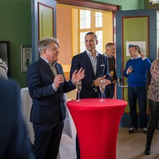 Zwei Männer im Anzug stehen vor einem roten Stehtisch mit Sektgläsern und prosten sich zu. Im Hintergrund halten weitere Personen ebenfalls Gläser, eine Frau mit Sektglas in der Hand. Die Szene wirkt wie eine kleine Feier oder Preisverleihung.