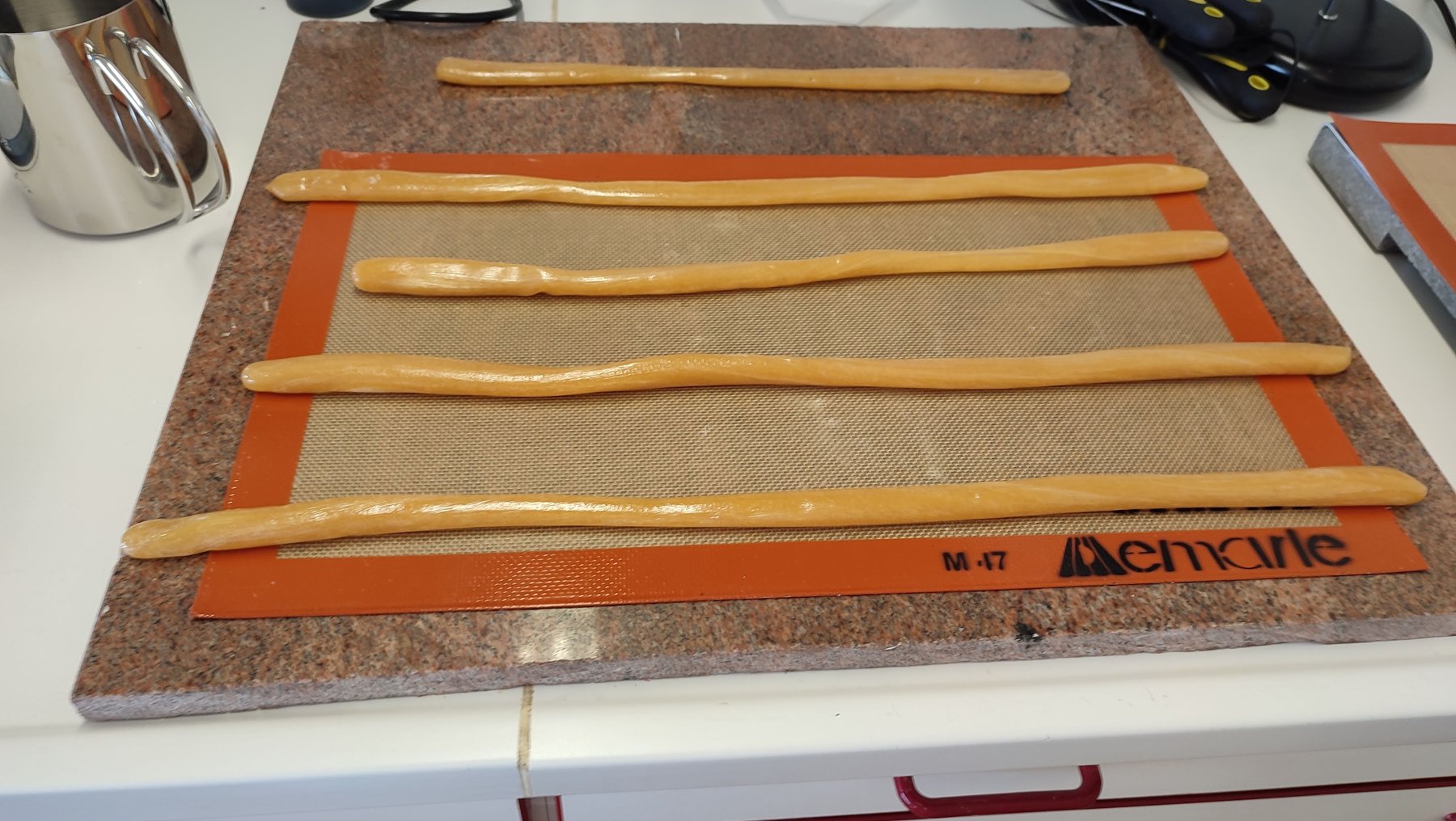 "Auf einem Backblech mit Backmatte liegen fünf längliche Teigstücke, die wie Brotstangen geformt sind. Die Backmatte hat eine orangefarbene Kante mit der Aufschrift 'Demarle'. Die Teigstücke sind gleichmäßig auf dem Blech verteilt."