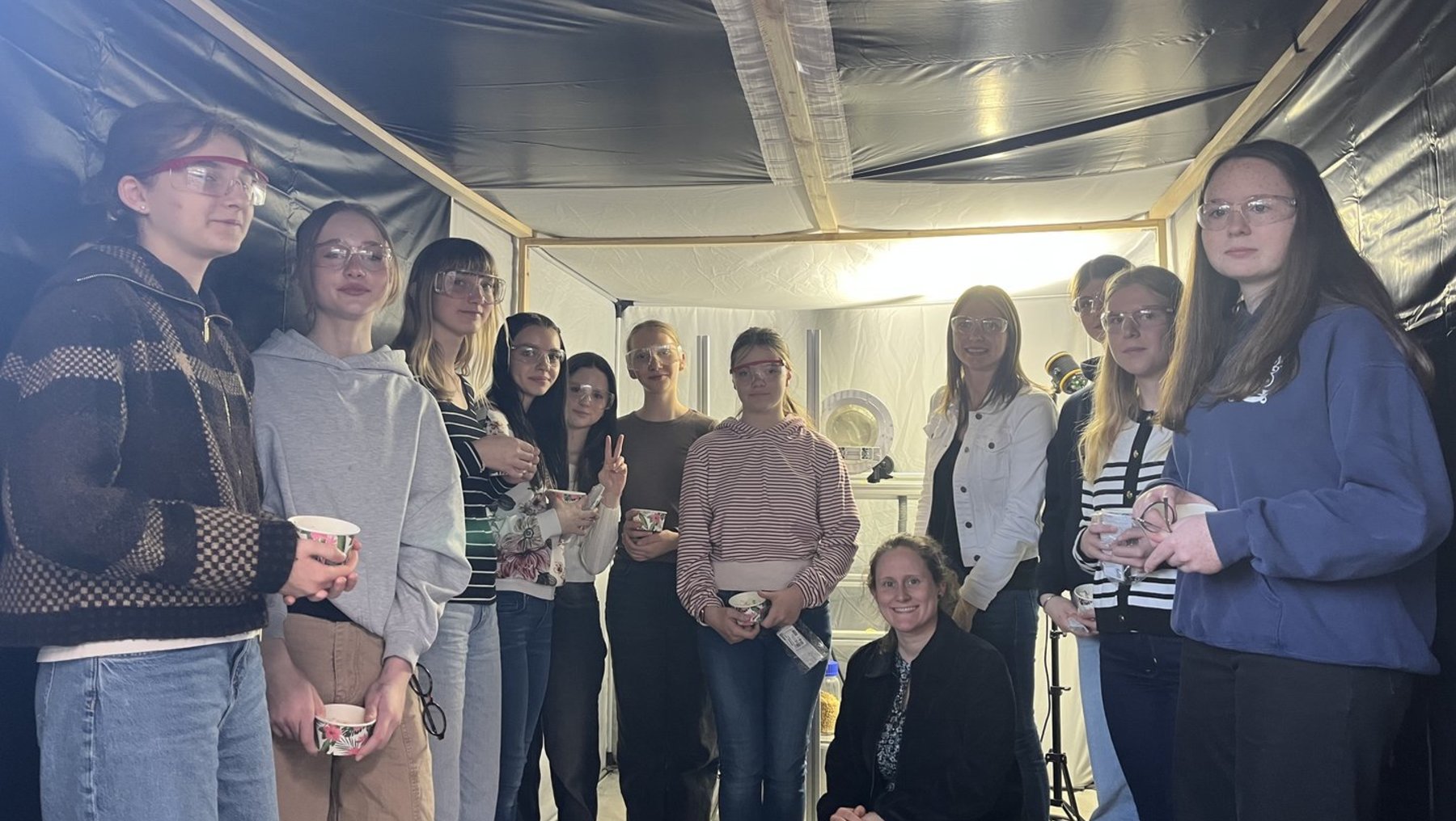 Gruppe von 14 Personen, die in einem Raum mit dunkler Decke und indirektem Licht stehen, halten Tassen und lächeln. Eine Person sitzt im Vordergrund, die anderen stehen in zwei Reihen. Gemütliche Atmosphäre mit lockerer Kleidung.