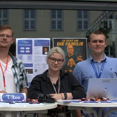 Drei Personen stehen hinter einem Tisch mit Laptops und Werbematerial. Sie lächeln in die Kamera. Im Hintergrund ist ein Poster mit dem Titel "55 Jahre um die Familie" und ein Gebäude zu sehen. Die Personen tragen Namensschilder und scheinen bei einer Veranstaltung zu sein.