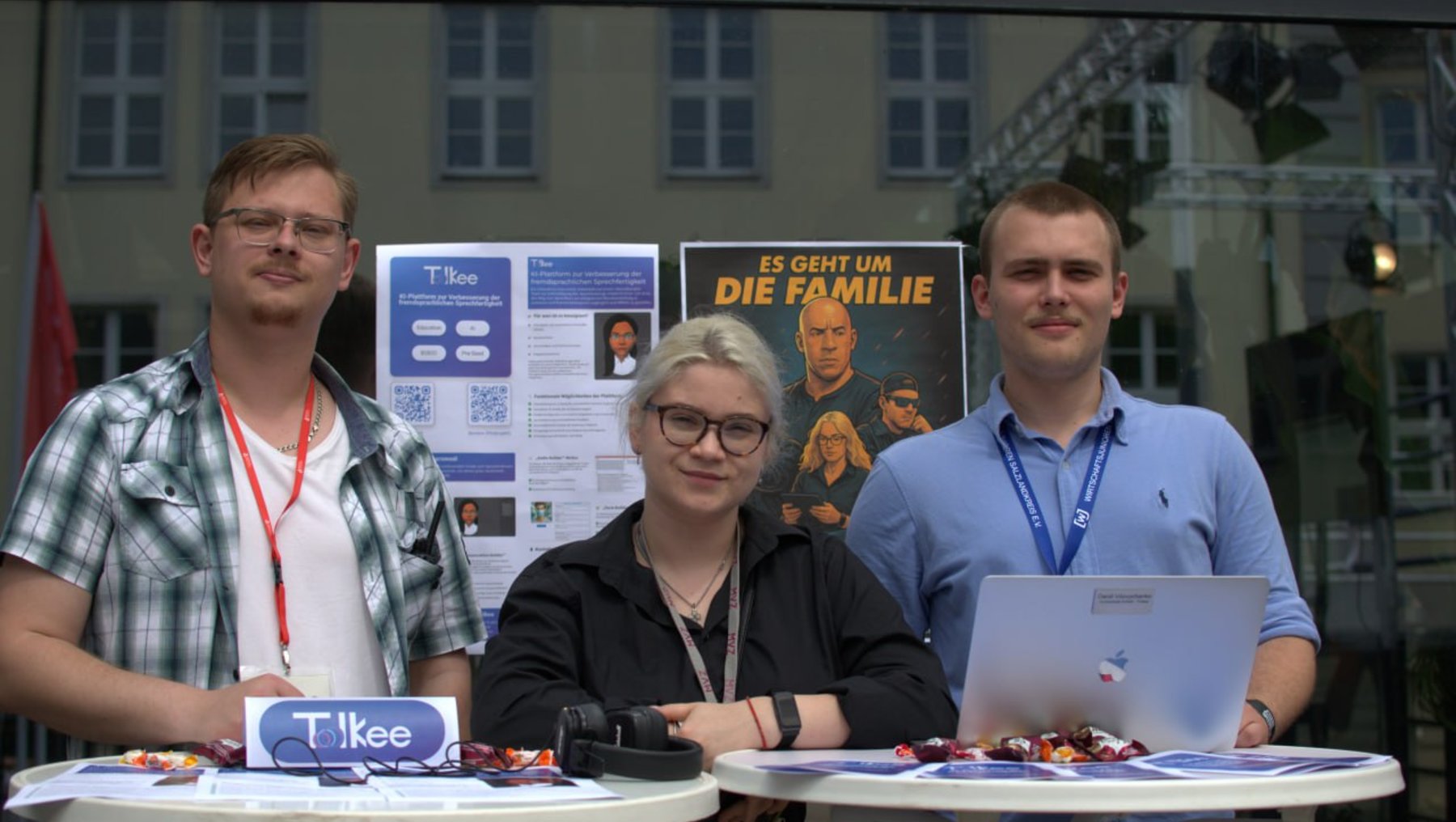 Drei Personen stehen hinter einem Tisch mit Laptops und Werbematerial. Sie lächeln in die Kamera. Im Hintergrund ist ein Poster mit dem Titel "55 Jahre um die Familie" und ein Gebäude zu sehen. Die Personen tragen Namensschilder und scheinen bei einer Veranstaltung zu sein.