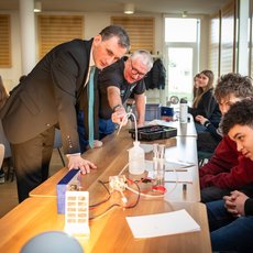 Zwei Männer stehen über einem Tisch und erklären einer Gruppe von Jugendlichen ein technisches Experiment mit Kabeln, einer Batterie und einem einfachen Schaltkreis. Die Jugendlichen beobachten aufmerksam. Sie experimentieren mit Wasserstoff. Zu Gast der Bildungsminister Sachsen-Anhalts Jan Riedel