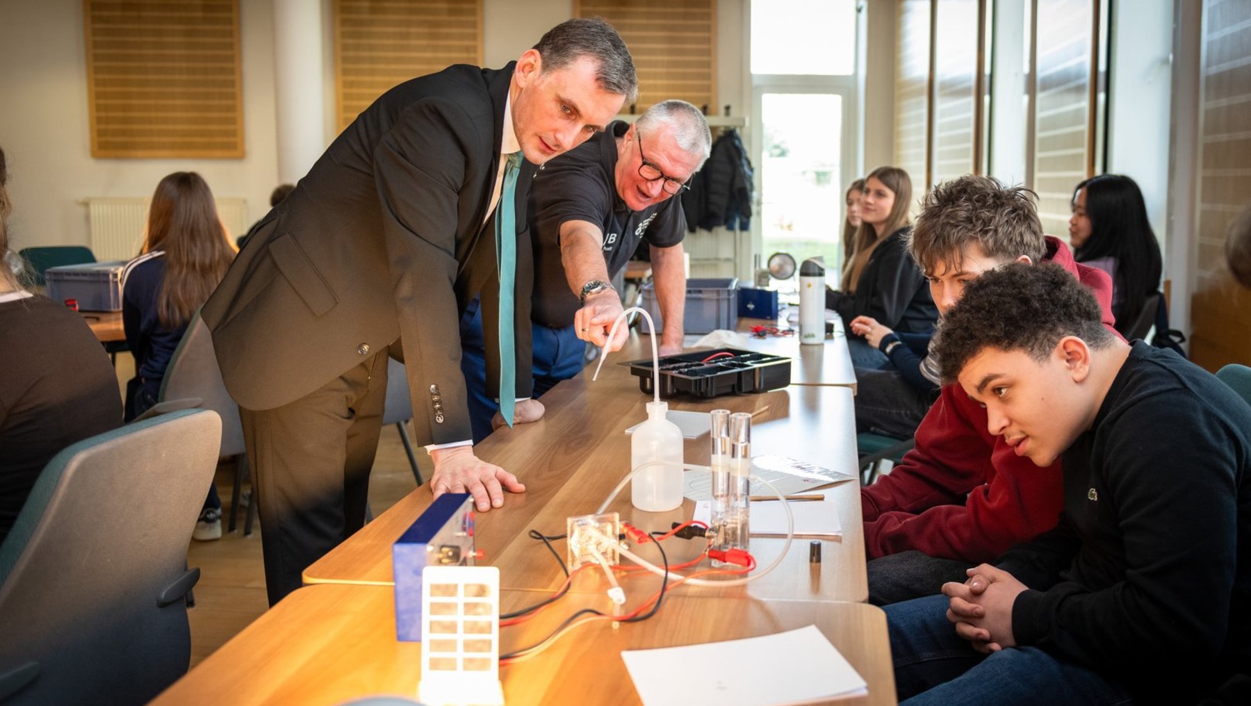 Zwei Männer stehen über einem Tisch und erklären einer Gruppe von Jugendlichen ein technisches Experiment mit Kabeln, einer Batterie und einem einfachen Schaltkreis. Die Jugendlichen beobachten aufmerksam. Sie experimentieren mit Wasserstoff. Zu Gast der Bildungsminister Sachsen-Anhalts Jan Riedel