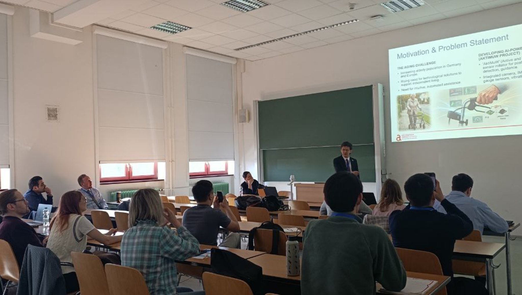 Ein Vortrag im Seminarraum: Ein Referent steht am Podium und präsentiert eine Folie mit dem Titel „Motivation & Problemstellung“, während etwa 20 Zuhörer auf Stühlen im Halbkreis sitzen und zuhören.