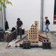Drei Personen bauen und gestalten eine große Modellstadt aus Holzbausteinen und anderen Materialien auf einem gepflasterten Boden. Im Vordergrund liegen weitere Bausteine und Werkzeuge bereit.