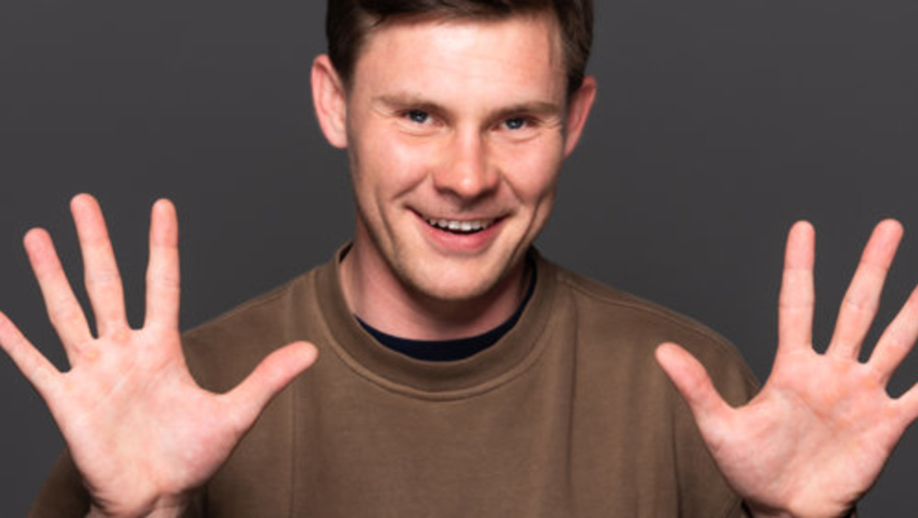 Ein lächelnder Mann mit hellen Haaren, der in einem braunen Adidas-Pullover vor dunklem Hintergrund steht und beide Hände mit gespreizten Fingern nach außen zeigt.