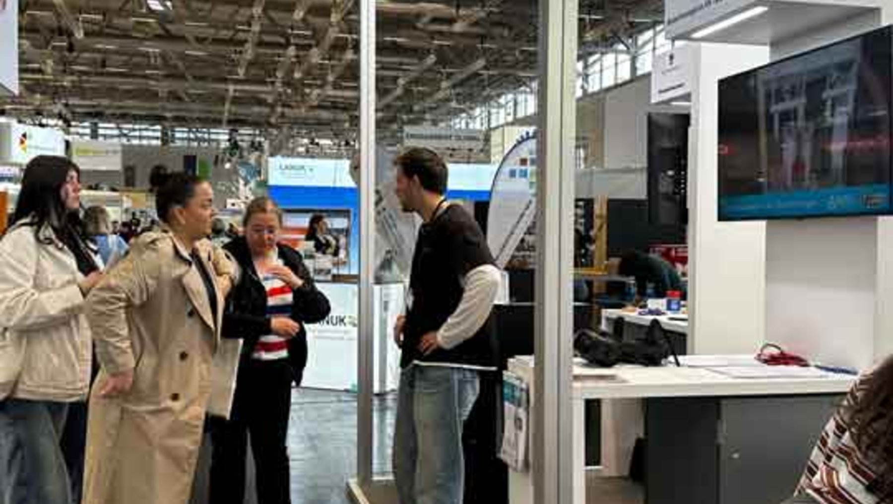 Eine Gruppe von fünf Personen steht auf einer Messe und unterhält sich vor einem Messestand mit einem großen Bildschirm, der eine Stadtansicht zeigt. Der Stand ist mit Infomaterial und einem Logo beschriftet.