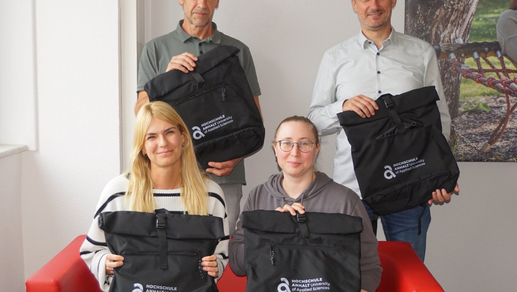 Vier Personen sitzen und stehen lächelnd zusammen, jede hält oder hält einen schwarzen Rucksack mit weißem Logo und Aufdruck „Innovationspreis für Klimaschutz“. Die Gruppe wirkt zufrieden und präsentiert die Rucksäcke als Auszeichnung.