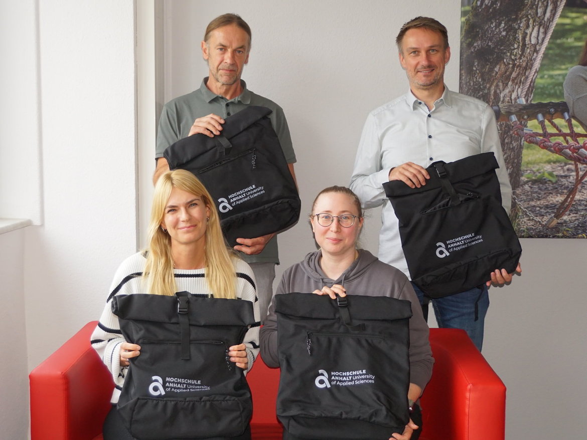 Vier Personen sitzen und stehen lächelnd zusammen, jede hält oder hält einen schwarzen Rucksack mit weißem Logo und Aufdruck „Innovationspreis für Klimaschutz“. Die Gruppe wirkt zufrieden und präsentiert die Rucksäcke als Auszeichnung.
