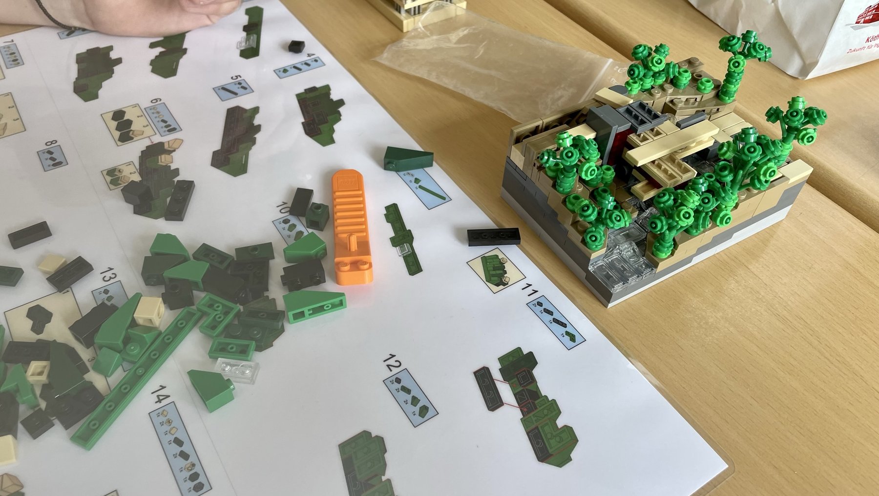 Eine Hand sortiert grüne und schwarze Lego-Steine auf einer Bauanleitung mit nummerierten Schritten. Im Hintergrund steht ein fertiges Modell eines kleinen Gebäudes mit grünen Bäumen und Details auf einer grauen Basis.