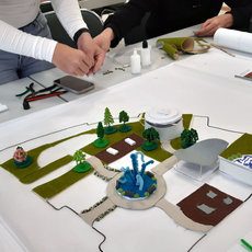 Zwei Personen arbeiten gemeinsam an einem großen Modell eines Stadt- oder Parkgeländes mit Gebäuden, Wegen, Grünflächen und einem zentralen Platz mit Brunnenanlage.