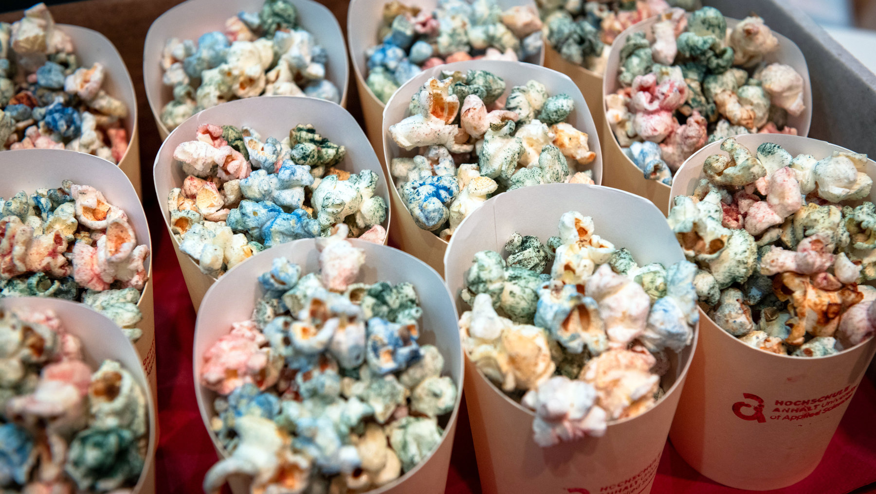 Mehrere Pappbecher mit buntem, gesalzenem Popcorn in verschiedenen Farben wie grün, blau, rosa und weiß, angeordnet auf einem Tisch.