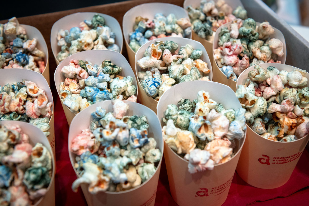 Mehrere Pappbecher mit buntem, gesalzenem Popcorn in verschiedenen Farben wie grün, blau, rosa und weiß, angeordnet auf einem Tisch.