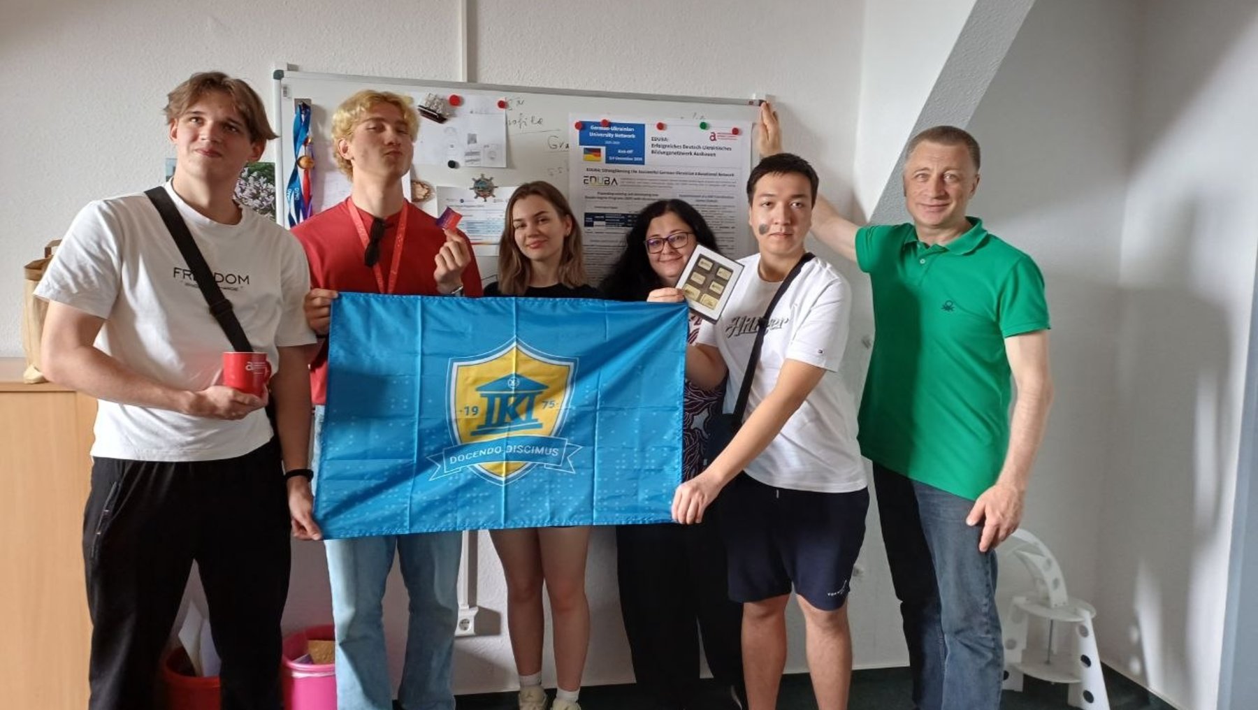 Fünf Personen halten gemeinsam eine blaue Fahne mit einem gelben Emblem und der Aufschrift „JK“. Dahinter hängt ein Poster mit Informationen und Symbolen zu einem Projekt oder einer Organisation.