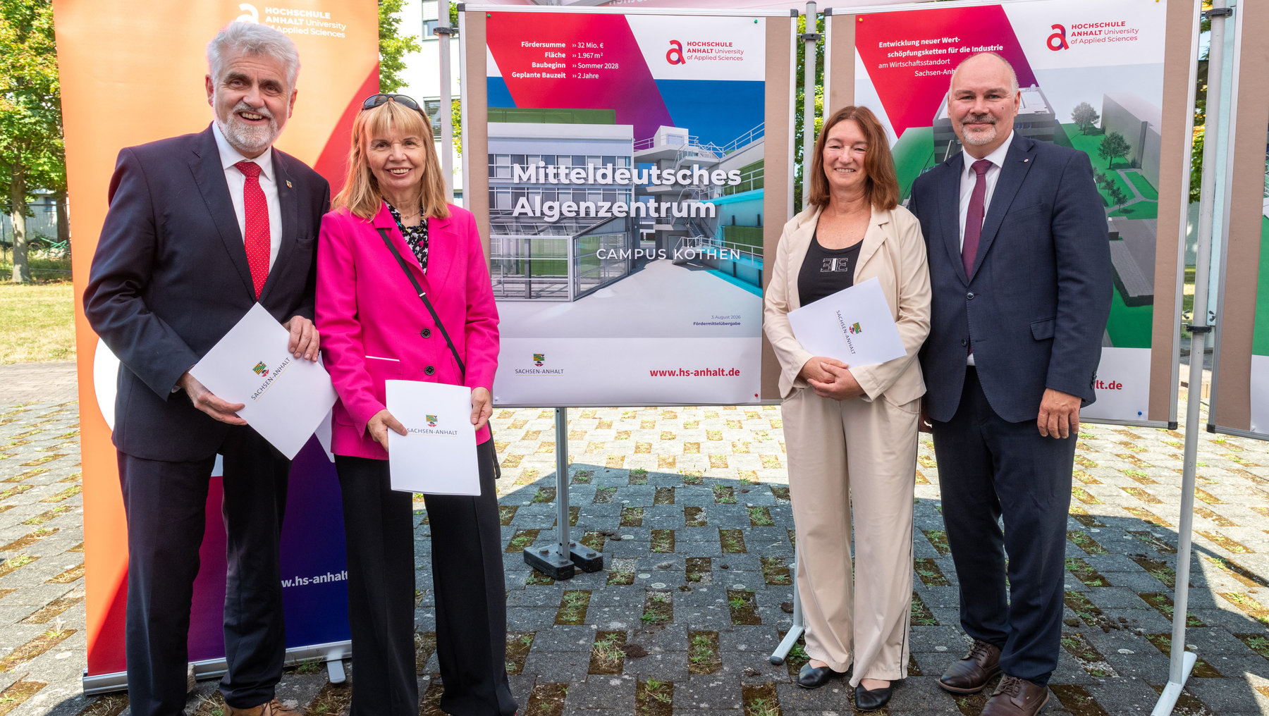 Vier Personen stehen vor einem Banner zur Eröffnung des „Mitteldeutschen Algenzentrums“ am Campus Kothen der Hochschule Anhalt. Sie halten Unterlagen und lächeln. Im Hintergrund ist ein Gebäudeentwurf mit Infos zu Förderung und Bauzeit zu sehen.