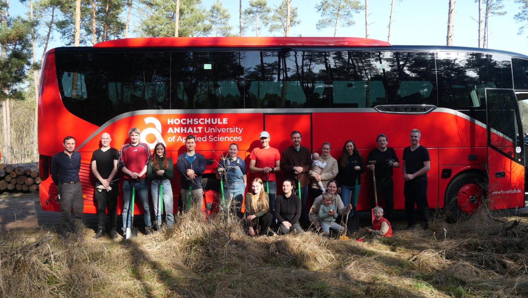 Gruppe von etwa 15 Personen, darunter Studierende und Betreuer, posiert lächelnd vor einem roten Reisebus der **Hochschule Anhalt – University of Applied Sciences**. Der Bus steht auf einem Feldweg vor einem Wald.