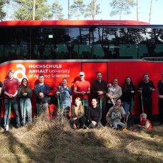 Gruppe von etwa 15 Personen, darunter Studierende und Betreuer, posiert lächelnd vor einem roten Reisebus der **Hochschule Anhalt – University of Applied Sciences**. Der Bus steht auf einem Feldweg vor einem Wald.