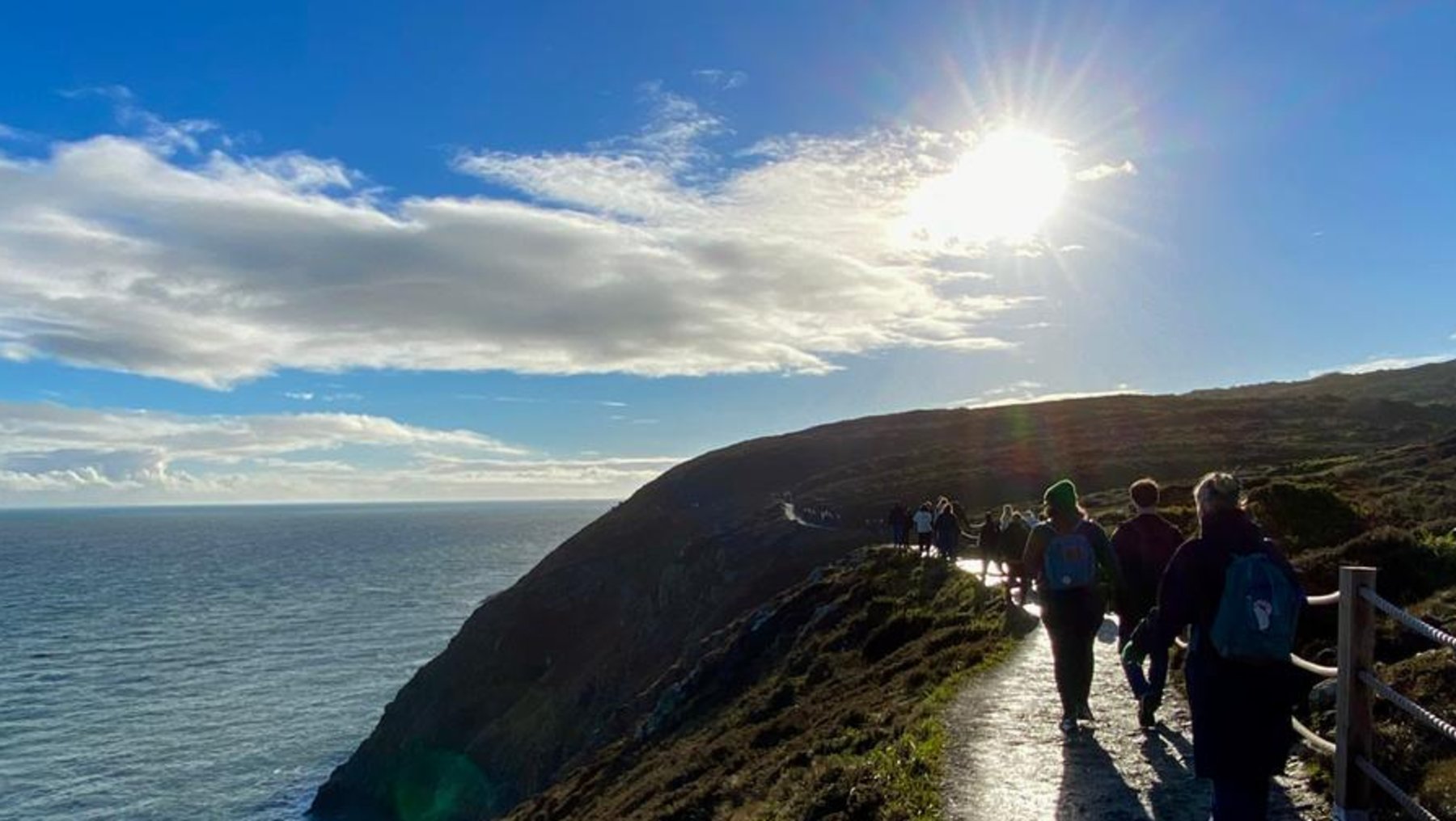 Ein sonniger Wanderweg führt entlang einer steilen Klippe mit Blick aufs Meer. Mehrere Personen gehen auf dem gepflasterten Pfad, der sich durch grüne Vegetation schlängelt. Die Sonne scheint hell am Himmel, während das Meer in der Ferne zu sehen ist.