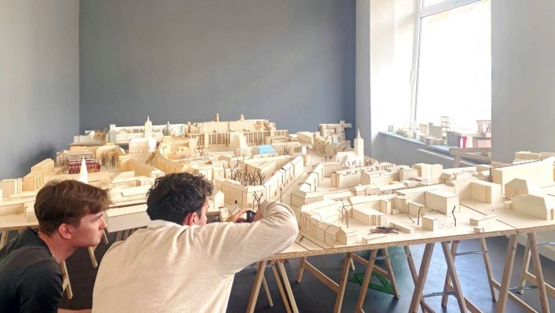 Zwei Personen betrachten ein detailliertes Modell einer Stadt aus Holz. Das Modell zeigt verschiedene Gebäude und Straßen in Miniaturform. Die Szene spielt in einem hellen Raum mit großen Fenstern. Die Personen scheinen das Modell zu analysieren oder zu besprechen.