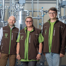 Drei Personen in Arbeitskleidung mit grünen Westen und einem Logo stehen vor großen Brauereikesseln und lächeln in die Kamera. Die Umgebung wirkt wie eine moderne Brauerei mit Edelstahlrohren und -behältern.