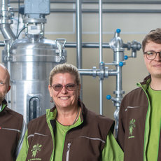 Drei Personen in Arbeitskleidung mit grünen Westen und einem Logo stehen vor großen Brauereikesseln und lächeln in die Kamera. Die Umgebung wirkt wie eine moderne Brauerei mit Edelstahlrohren und -behältern.