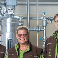 Drei Personen in Arbeitskleidung mit grünen Westen und einem Logo stehen vor großen Brauereikesseln und lächeln in die Kamera. Die Umgebung wirkt wie eine moderne Brauerei mit Edelstahlrohren und -behältern.