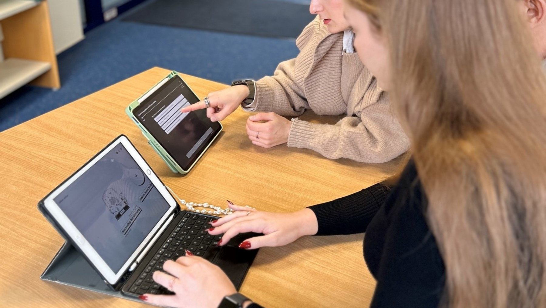 Zwei Personen sitzen an einem Tisch und arbeiten gemeinsam an Laptops. Eine Person zeigt etwas auf dem Bildschirm des anderen Laptops. Beide wirken konzentriert und engagiert. Der Raum ist hell und modern eingerichtet.