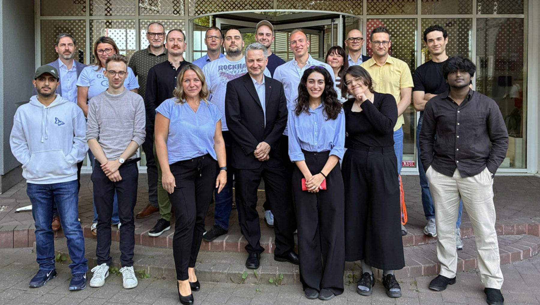 Gruppe von 16 Personen, darunter Männer und Frauen, steht lächelnd vor einem Gebäude. Die meisten tragen blaue Shirts, einige haben andere Kleidung. Sie wirken zusammengehörig und professionell.