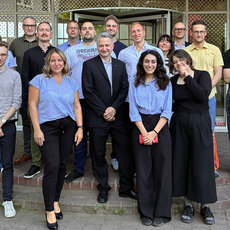 Gruppe von 16 Personen, darunter Männer und Frauen, steht lächelnd vor einem Gebäude. Die meisten tragen blaue Shirts, einige haben andere Kleidung. Sie wirken zusammengehörig und professionell.